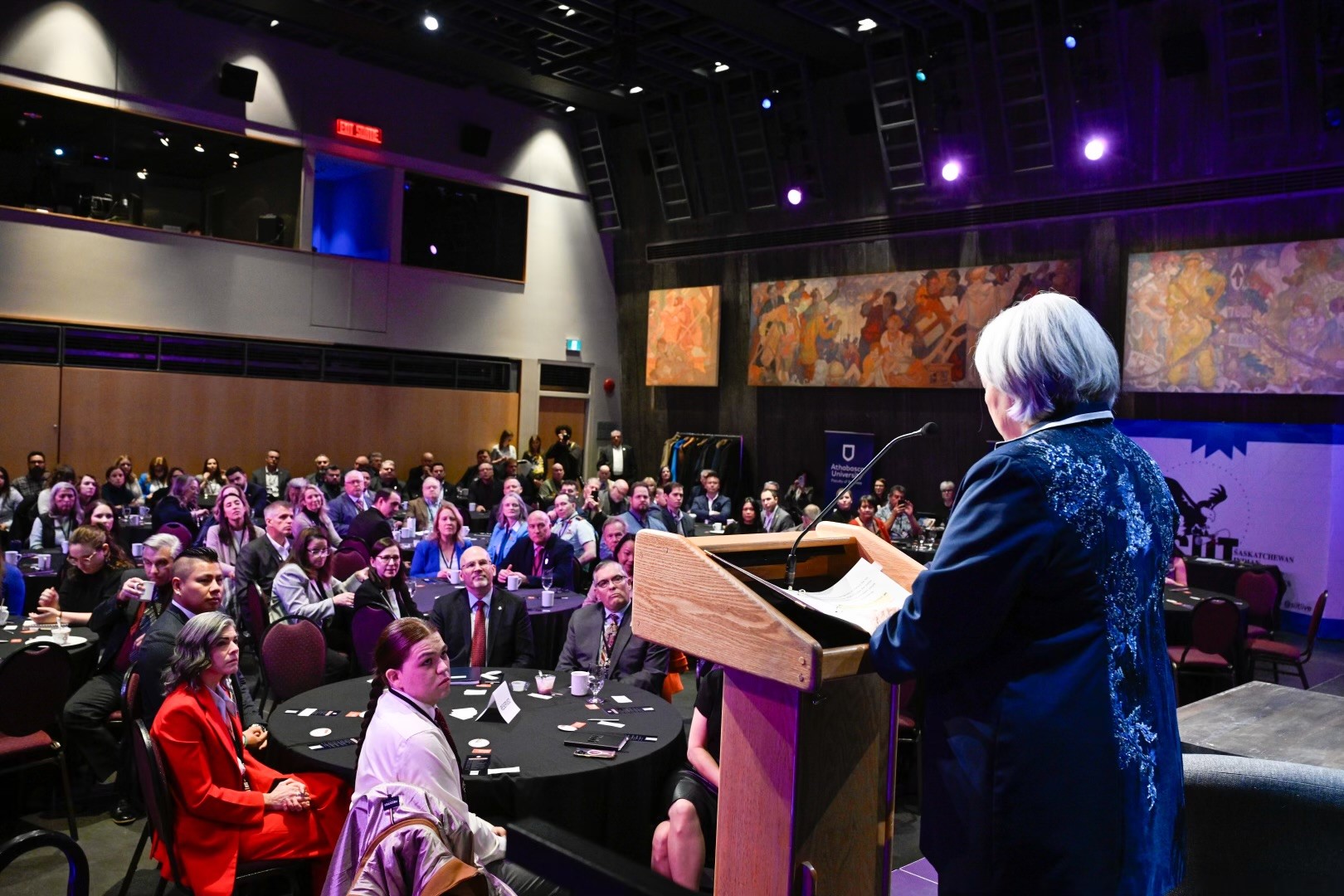 La gouverneure générale Mary Simon est vue de dos, debout, parlant à un podium, sur une scène, devant une salle remplie de personnes assises à des tables rondes.