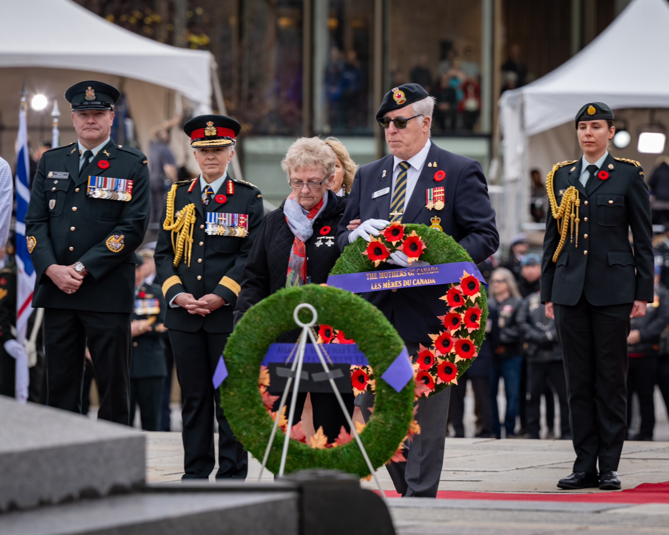 A military member with grey hair is holding a wreath of flowers with a ribbon that says "The Mothers of Canada". A lady with glasses, wearing a light blue and red scarf around her neck, bowing her head, is next to him. they are surronded by miltrary membe