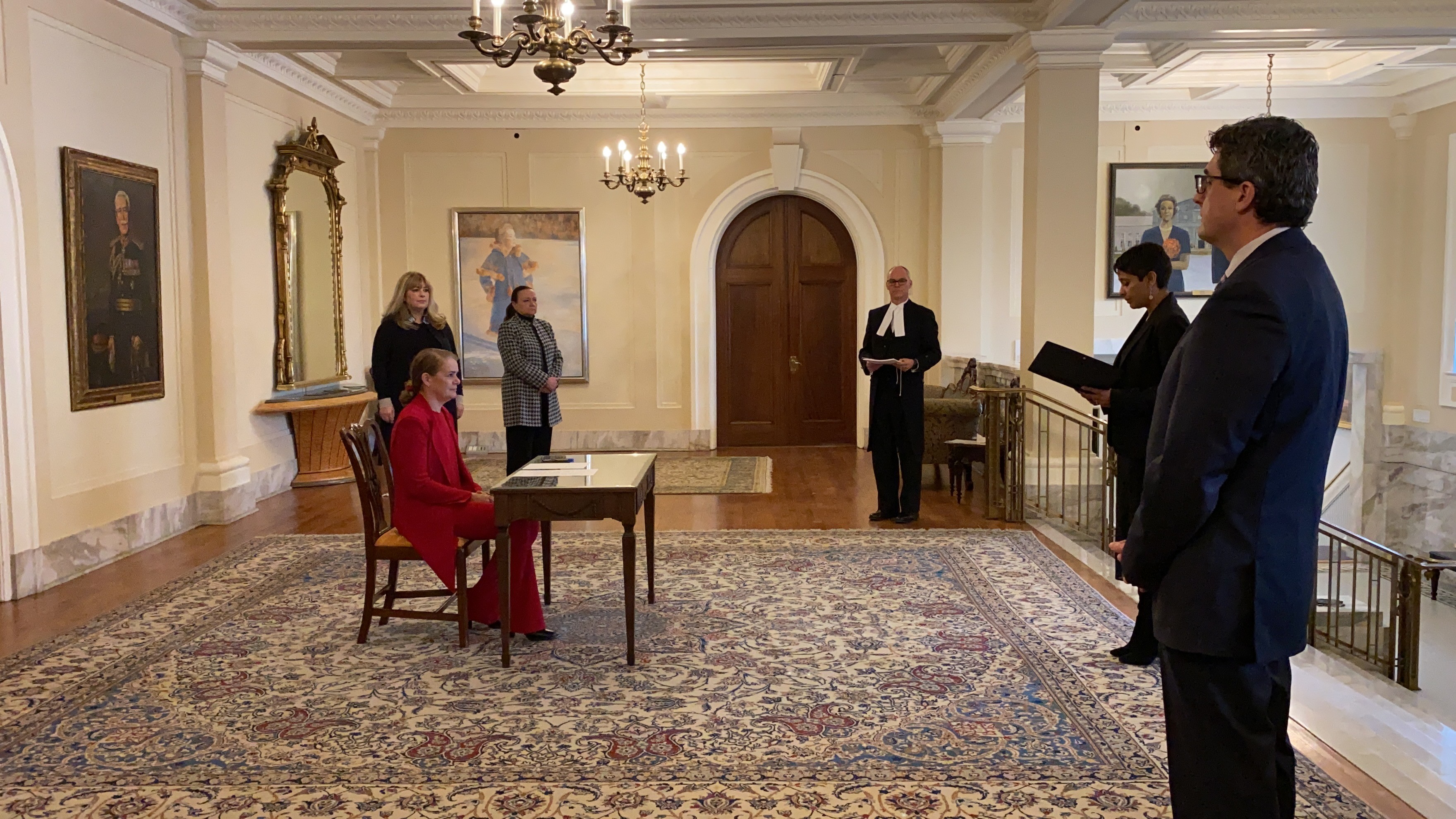 The Governor General is sitting at a desk. On the right hand side, there is a man holding a document in his hands. Four other people are standing around the desk all while respecting the social distancing norms.