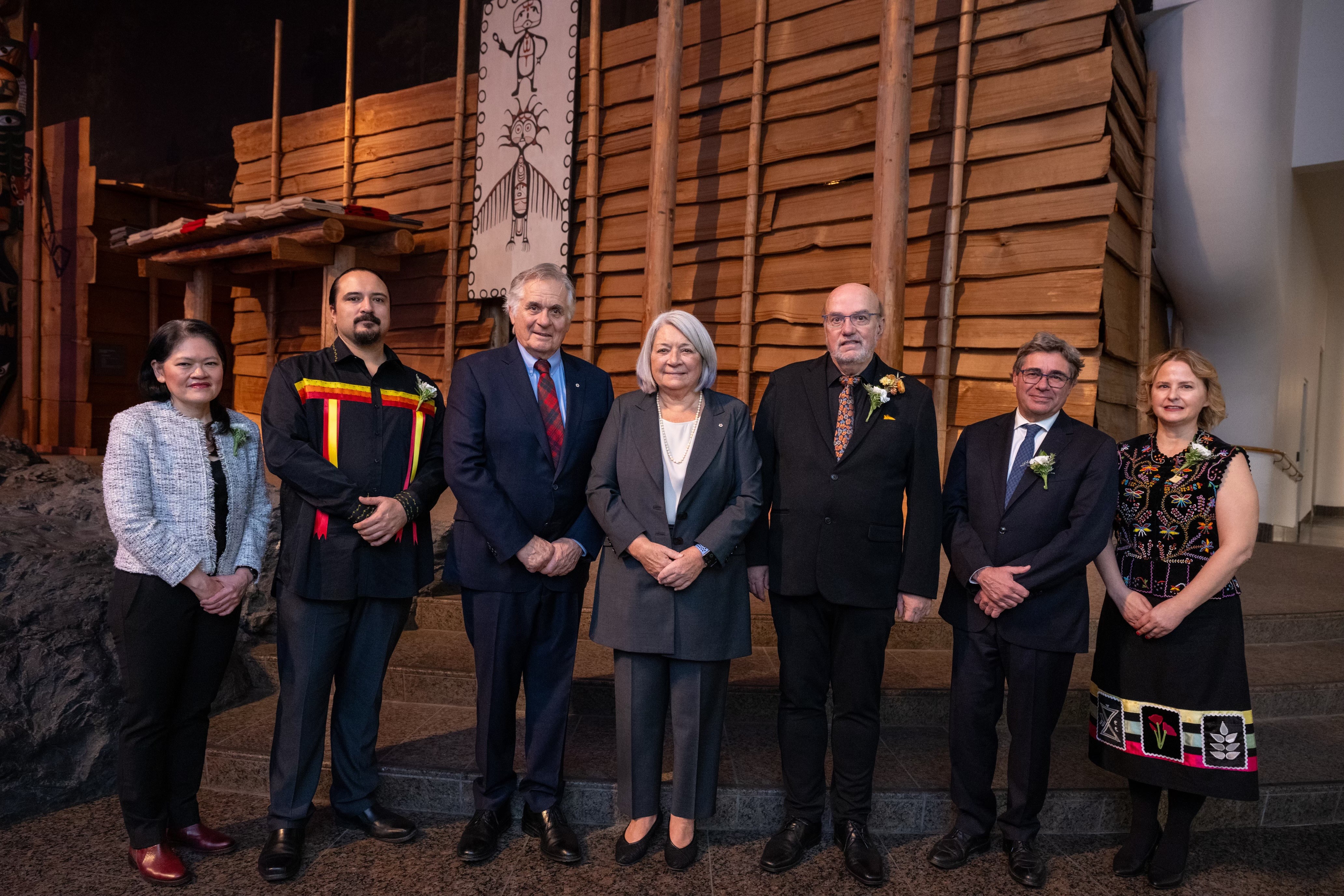 La gouverneure générale Mary Simon et M. Whit Fraser se tiennent au centre d'une file de 5 autres personnes, devant un mur en bois. Tous sourient à la caméra.