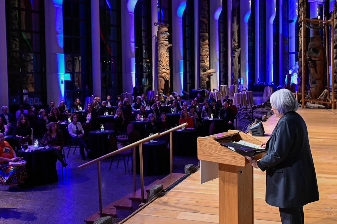 La gouverneure générale Mary Simon est vue du côté gauche, s'exprimant à un podium, devant une foule de personnes assises à des tables rondes. Des lumières violettes et bleu clair sont projetées sur de hauts piliers derrière le public.