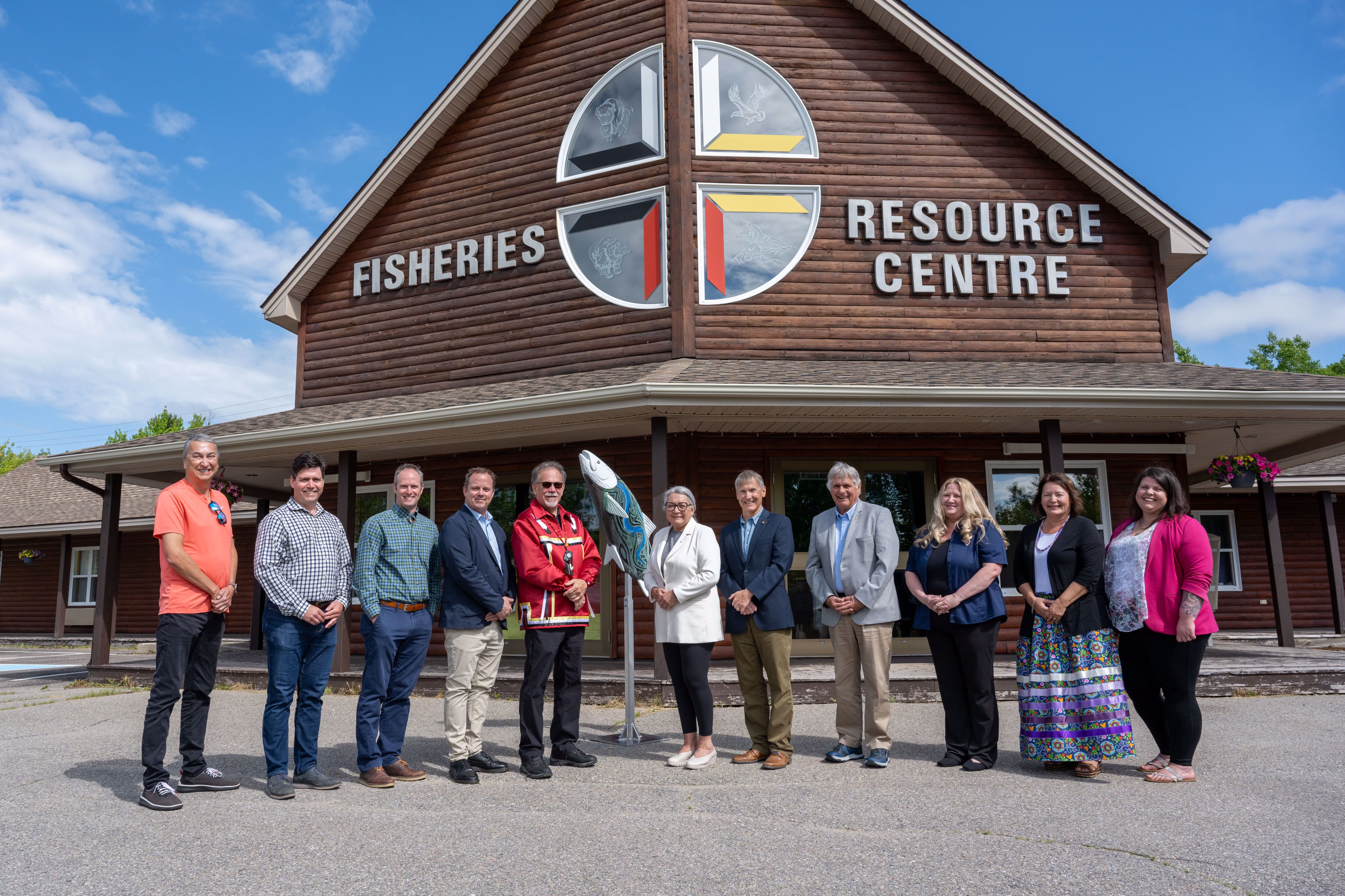 La gouverneure générale Simon et M. Whit Fraser se tiennent à l'extérieur avec un groupe de personnes. Un grand bâtiment avec les mots « Fisheries Resource Centre » sur la façade se trouve derrière eux.