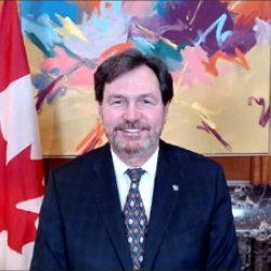 Screenshot of the Administrator smiling at the camera. There is a Canadian flag to his left at a brightly coloured painting behind him.
