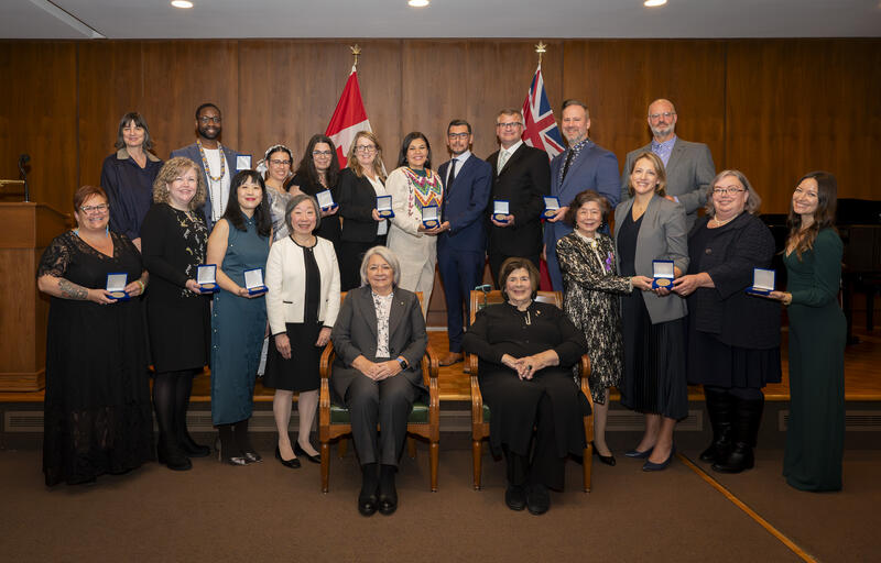 A group of 18 men and women are posing for the camera, standing and holding open boxes containing a medallion. Governor General Mary Simon is seated in a chair, in the centre, with a lady sitting next to her.]