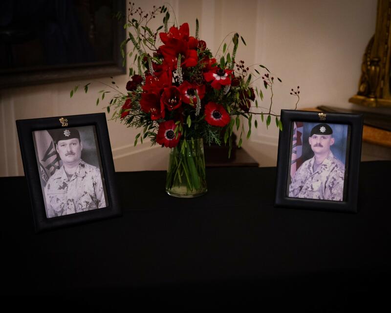 Two photos of military members are showcased on a table on each sides of a vase holding red flowers. The table is covered by a black tablecloth.