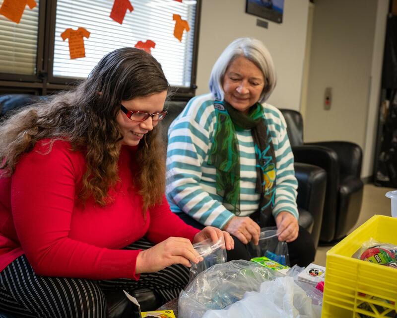 Governor General Mary Simon, seated, wearing a striped white and green sweater with a green scarf is looking at a plastic bag presented by a lady wearing red glasses and a red sweater.