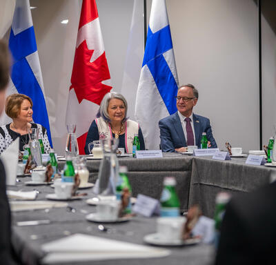 La gouverneure générale Mary Simon est assise à la tête d’une grande table de conférence, entre deux autres personnes. Derrière elle se trouvent deux drapeaux du Canada et deux drapeaux de la Finlande.