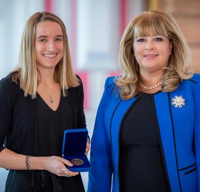 À gauche, une étudiante universitaire blonde tient une boîte bleue ouverte contenant une médaille. À droite, une femme blonde portant une veste bleue.