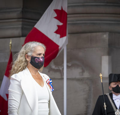 Une femme vêtue d'un costume blanc avec un masque noir est en train de marcher. Un homme en uniforme noir et un agent de la Gendarmerie royale du Canda portant un uniforme rouge saluent en arrière-plan. Il y a également deux drapeaux canadiens.