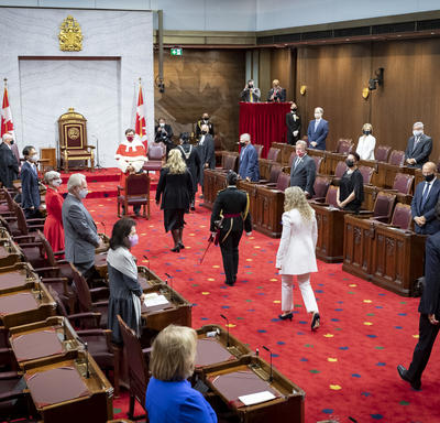 Un cortège de personnes marche sur un tapis rouge vers l'avant d'une grande salle au tapis rouge. Trois des personnes sont vêtues de noir. Une femme est vêtue de blanc. Il y a un trône flanqué de deux drapeaux du Canada à l'avant de la salle.