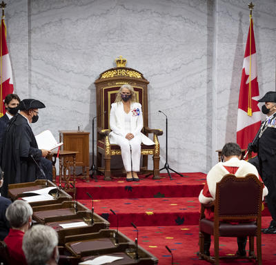 Une femme vêtue d'un costume blanc est assise sur un trône. Il y a des drapeaux du Canada de chaque côté de la plate-forme. Un homme assis sur une chaise lui fait face. Il porte un manteau rouge et blanc.