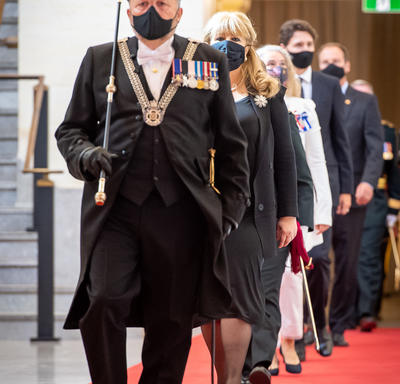 A line of people are walking towards the camera on a red carpet. The man leading the group is wearing a decorated black uniform, carrying a black and gold rod. He is also wearing a black cap and a black face mask.