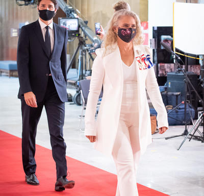 A woman in a white pant suit is walking on a red carpet. She is wearing black heels and a black face mask. A man is walking behind her in a dark suit, also wearing a face mask.