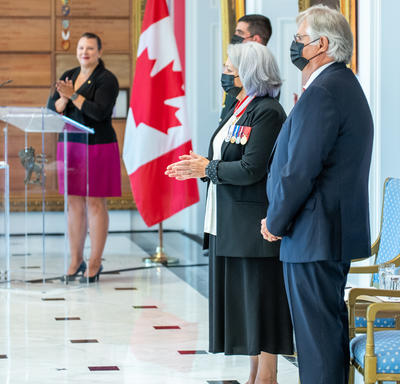 Governor General Mary May Simon claps. His Excellency Whit Fraser stands with hands folded in front of him. A Canadian flag and the emcee of the event are in the background.