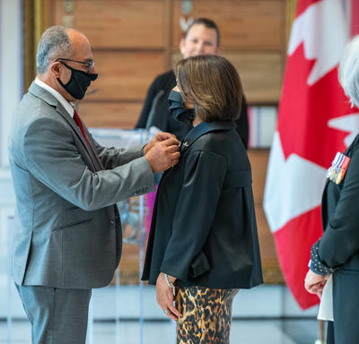 Un homme épingle une distinction sur le blouson d’une femme, sous le regard de la gouverneure générale Mary May Simon. Un drapeau canadien décore le fond de la pièce.