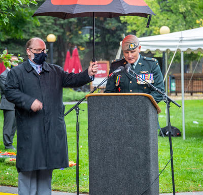 Un homme en uniforme parle derrière un lutrin. Un autre homme tient un parapluie pour le protéger de la pluie.