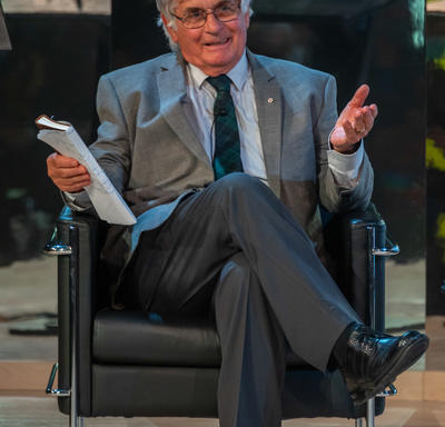 Mr.  Fraser is sitting with his legs crossed in a black leather chair. He has a notebook in his right hand with a piece of paper wrapped around it. His left hand is open and raised to the side in a relaxed gesture. He is smiling.