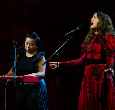 Iskwē et une musicienne accompagnatrice sont sur une scène. Iskwē porte une robe rouge et chante dans un microphone. La musicienne accompagnatrice porte une robe noire et est assise derrière un clavier en train de jouer une chanson.