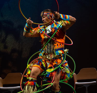 Dallas Arcand is on a stage, he is performing an Indigenous hoop dance with 11 hoops. He is dressed in a traditional outfit that is very colourful. Behind him are several wooden chairs. 