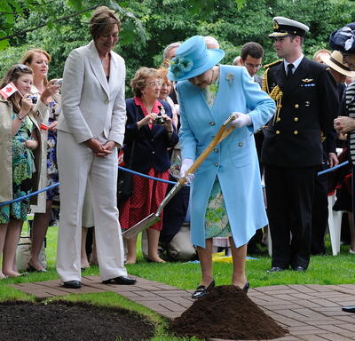 The Queen, in a matching blue coat and hat, shovels soil during a tree-planting ceremony, as a crowd of people look on. Stands of trees are in the background