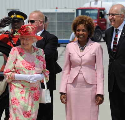 La Reine est debout aux côtés de la gouverneure générale Michaëlle Jean et de Jean-Daniel Lafond. Ils sont à l’extérieur d’un aéroport. Un agent de la GRC et du personnel de sécurité sont derrière eux.