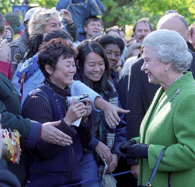 The Queen, in a bright green coat, smiles at a crowd of people standing outside. Several people are reaching forward to shake The Queen’s hand.