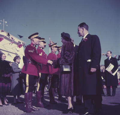 Princess Elizabeth, wearing a fur coat, shakes hands with an RCMP officer while three other officers stand at attention. The officers are wearing red dress uniforms. A ship and nautical flags are in the background.