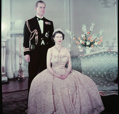The Queen Elizabeth and The Duke of Edinburgh, both wearing formal attire, pose for a portrait. The Queen is seated on a light blue chair. A floral arrangement is on a table behind her. 