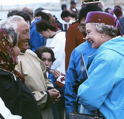 La Reine, qui porte un parka bleu et un chapeau rouge bordeaux, sourit à deux Aînés inuits en habits traditionnels. Un enfant tenant un petit drapeau du Canada regarde la scène. On voit une foule à l’arrière-plan.