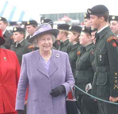La Reine, qui porte un manteau mauve et un chapeau assorti, passe devant un groupe de cadets en uniforme au garde-à-vous.