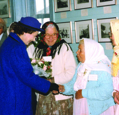 La Reine serre la main d’une femme portant un fichu sous le regard de deux autres femmes à la tenue similaire.