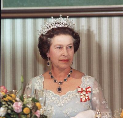 The Queen, wearing a formal dress, the Order of Canada insignia and a jeweled tiara, sits at a table. The table is draped with cloth, and a flower arrangement is in the foreground. 