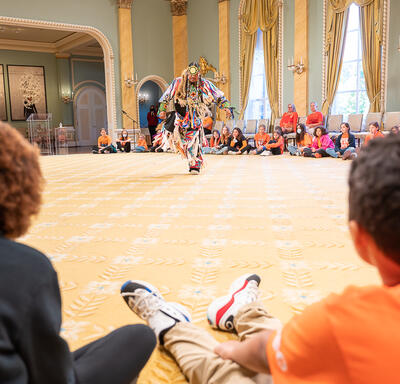 Un danseur se produit dans la salle de bal de Rideau Hall. Il porte une tenue traditionnelle. Des écoliers sont assis autour de lui et le regardent.