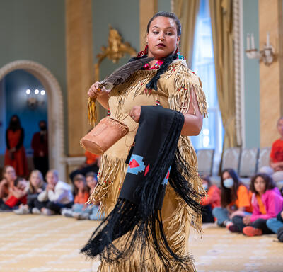 A woman is standing in the Ballroom at Rideau Hall. She is wearing a traditional Indigenous outfit and is holding a feather fan in front of her chest.