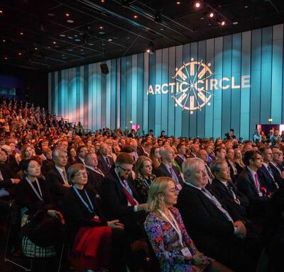 A large seated audience. The wall next to the audience is lit with blue lights and a projection on the wall reads, “Arctic Circle.”
