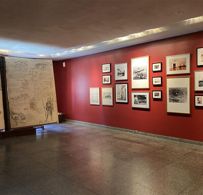Il y a une grande exposition de 2 pièces de croquis et de textes sur la gauche. Sur la droite, un mur rouge présente un mélange de 17 photographies, croquis et peintures.