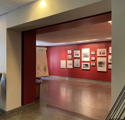 Vue à travers une large porte dans une pièce au mur rouge qui contient un mélange de photographies, de croquis et de peintures.