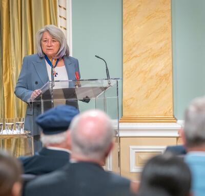 Governor General Simon stands in the Ballroom at Rideau Hall speaking into a microphone. There is an audience seated in front of her. 