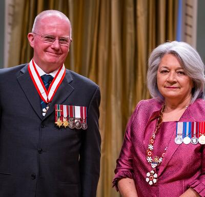 Governor General Simon is standing next to a man who has a medal hanging around his neck.