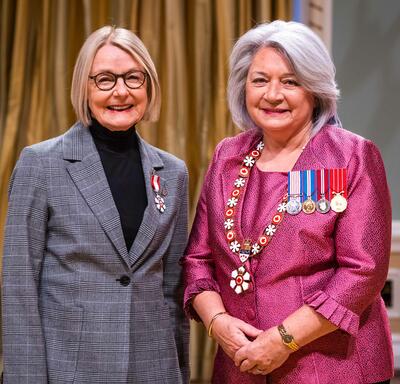 La gouverneure générale Simon se tient à côté d’une femme qui porte une médaille épinglée à son blazer gris.