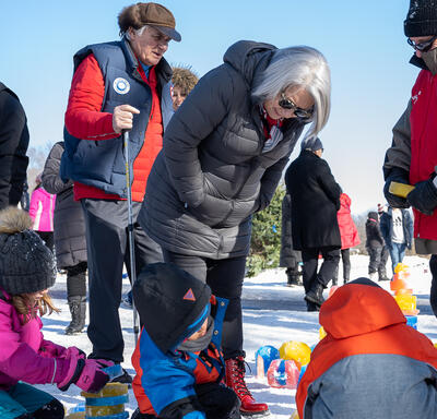 La gouverneure générale Mary Simon se penche pour parler à des enfants qui jouent dans la neige. Derrière elle se trouvent M. Fraser et plusieurs autres personnes.