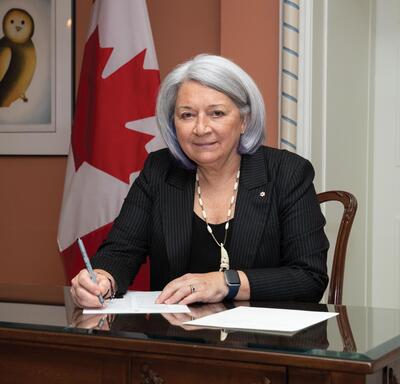 La gouverneure générale Mary Simon, assise à un bureau, signe un document papier. Un drapeau canadien est exposé derrière elle.