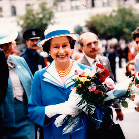 La reine Elizabeth II marche à l’extérieur. Elle porte un habit bleu et blanc et tient un bouquet de fleurs. La foule la regarde.
