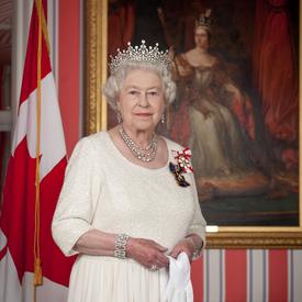 La reine Elizabeth II porte une robe de couleur crème et est coiffée d’une couronne. Elle tient une paire de gants blancs. Elle se tient devant un portrait de la reine Victoria. Un drapeau du Canada se trouve à l’arrière-plan.