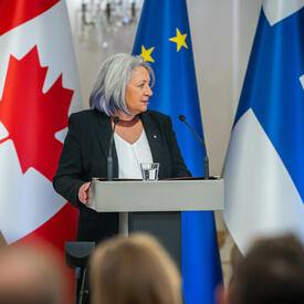 La gouverneure générale Mary Simon parlant dans un microphone, derrière un pupitre. Derrière elle se trouvent, de gauche à droite, le drapeau du Canada, le drapeau de la Finlande et le drapeau de l’Union européenne.