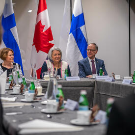 La gouverneure générale Mary Simon est assise à la tête d’une grande table de conférence, entre deux autres personnes. Derrière elle se trouvent deux drapeaux du Canada et deux drapeaux de la Finlande.