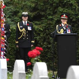 La gouverneure générale du Canada, en uniforme de l'armée canadienne, prend la parole à un podium, dans un cimetière, lors d'une commémoration du jour du Souvenir en Belgique. Son aide de camp féminin se tient à sa droite.