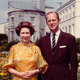 La Reine et le duc d’Édimbourg, les deux le sourire aux lèvres, se tiennent dans les jardins privés de Rideau Hall. La Reine porte une robe à motifs jaunes et oranges; le duc, un costume foncé.