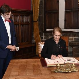 La gouverneure générale signe un livre d'or. Un grand homme se tient à sa droite et la surveille.