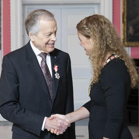 La gouverneure générale, Julie Payette, se tient debout à côté de Howard Gimbel. Ils se serrent la main. Tous deux portent l'insigne de l'Ordre du Canada.